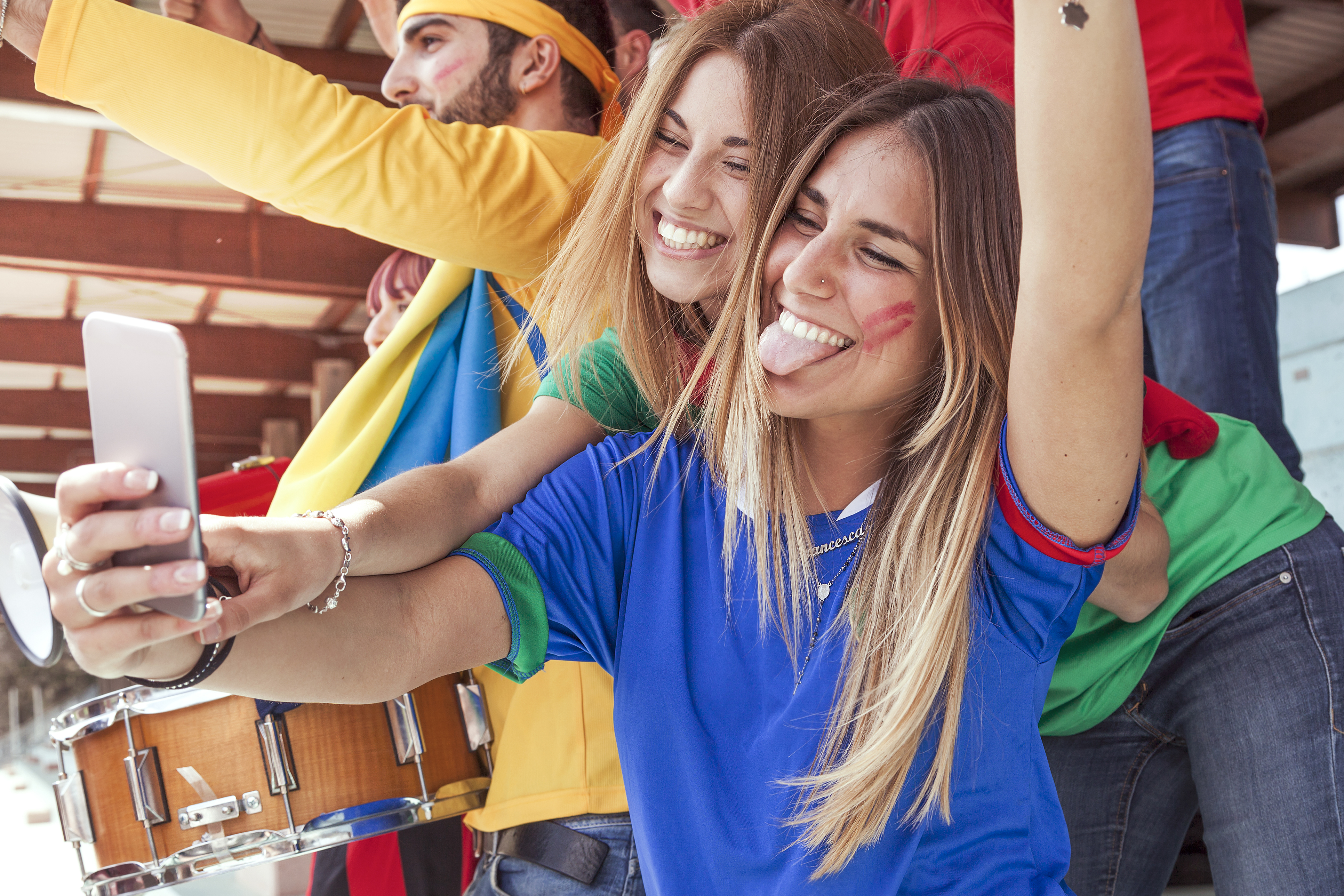 Zwei Freundinnen machen ein Selfie. Im Hintergrund sind mehrere Menschen zu erkennen, die alle bunt gekleidet sind. 