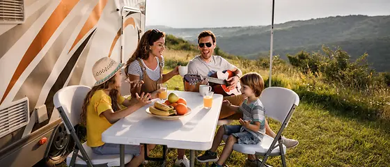 Familie beim Campen - Camping Urlaub Plus Europa Mutter, Vater und ihre zwei Kinder sitzen an einem Campingtisch und singen.