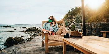 Eine Frau liegt auf einer Liege und trägt eine grüne Maske. Im Hintergrund sieht man das Meer.