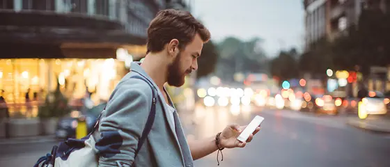 Ein Mann in der Stadt - Langzeit-Auslandskrankenversicherung bis 5 Jahre Ein Mann steht mit einem Rucksack in einer Stadt und guckt auf sein Handy.