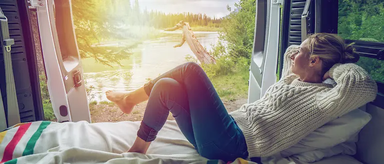 Eine Frau liegt in einem Camper. Im Hintergrund ist ein See zu sehen. 