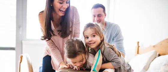 Familie mit Koffer - Gepäckversicherung Mutter, Vater und deren zwei Töchter sitzen auf einem Bett und probieren ihren Koffer zu zu bekommen.