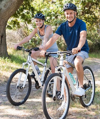 Zwei Personen sitzen auf dem Fahrrad und lächeln in die Kamera. Im Hintergrund ist ein sandiger Weg zu sehen.