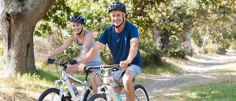 Zwei Personen sitzen auf dem Fahrrad und lächeln in die Kamera. Im Hintergrund ist ein sandiger Weg zu sehen.