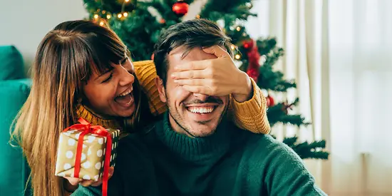 Eine Frau übergibt ihrem Mann ein Geschenk. Im Hintergrund ist ein geschmückter Weihnachtsbaum.