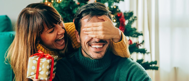 Eine Frau übergibt ihrem Mann ein Geschenk. Im Hintergrund ist ein geschmückter Weihnachtsbaum.