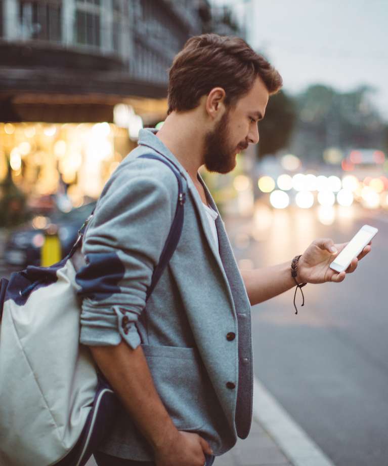 Ein Mann steht mit einem Handy in der Hand an einer Straße. Im Hintergrund sind Geschäfte zu sehen. 