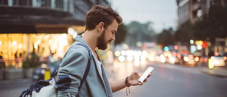 Ein Mann steht mit einem Handy in der Hand an einer Straße. Im Hintergrund sind Geschäfte zu sehen. 