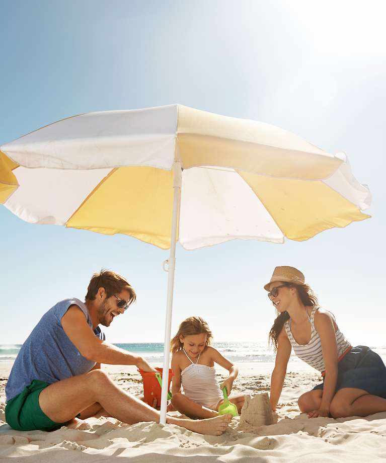 Eine Familie sitzt unter einem Sonnenschirm am Strand und spielen im Sand.