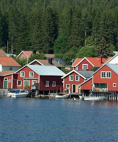 Kleine rote Häuser stehen am Wasser. Im Hintergrund ist ein Wald. 