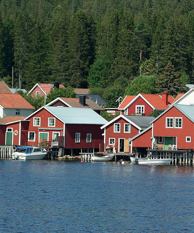 Kleine rote Häuser stehen am Wasser. Im Hintergrund ist ein Wald. 