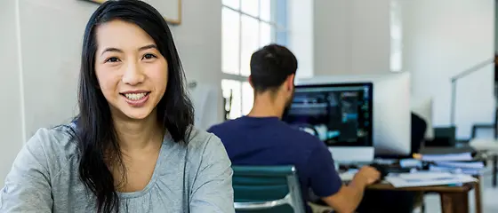 Zwei Personen am Arbeitsplatz - Hanse-Global Zwei Personen sitzen am Arbeitsplatz. Im Vordergrund sitzt eine Frau am Handy und schaut in Kamera.