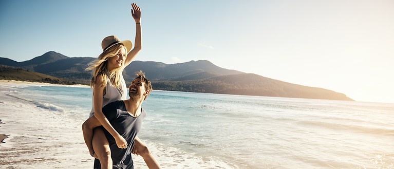 Ein Mann und eine Frau sind am Strand. Im Hintergrund kann man Wasser und grüne Hügel sehen. 