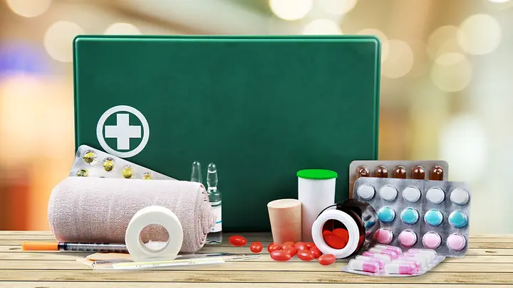 Im Vordergrund ist ein grüner Erste Hilfe Kasten zu sehen. Vor dem Kasten liegen Bandagen, verschiedene Tabletten und Pflaster. 