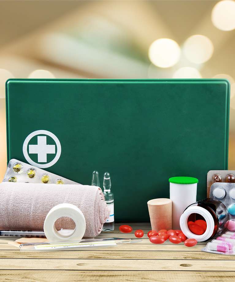 Im Vordergrund ist ein grüner Erste Hilfe Kasten zu sehen. Vor dem Kasten liegen Bandagen, verschiedene Tabletten und Pflaster. 