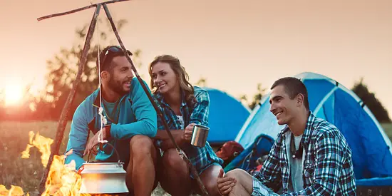Drei Freunde sitzen an einem Lagerfeuer und kochen. Im Hintergrund kann man zwei Zelte sehen. 