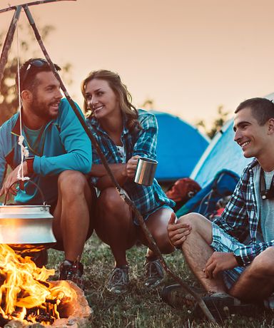 Drei Freunde sitzen an einem Lagerfeuer und kochen. Im Hintergrund kann man zwei Zelte sehen. 