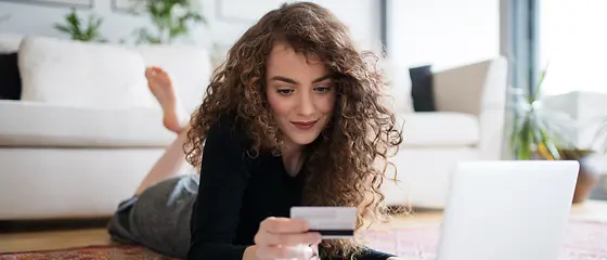 Eine Frau liegt auf dem Boden. Vor ihr steht ein Laptop. In ihrer Hand hat sie eine Kreditkarte. 