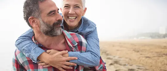 Eine Mann und eine Frau sind am Strand. Der Mann nimmt seine Frau Huckepack
