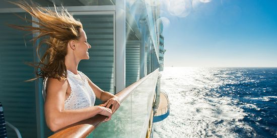Eine Frau steht an der Reling eines Kreuzfahrtschiffs und genießt den Wind und die Sonne. 