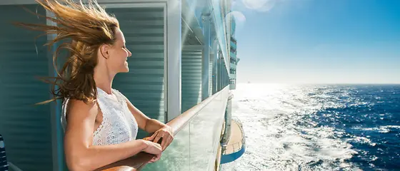 Eine Frau steht an der Reling eines Kreuzfahrtschiffs und genießt den Wind und die Sonne. 