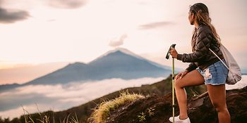 Eine Frau steht auf einem Berg und genießt die Aussicht. Im Hintergrund kann man einen anderen Berg und Wolken sehen. 