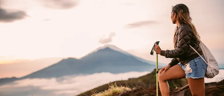 Eine Frau steht auf einem Berg und genießt die Aussicht. Im Hintergrund kann man einen anderen Berg und Wolken sehen. 