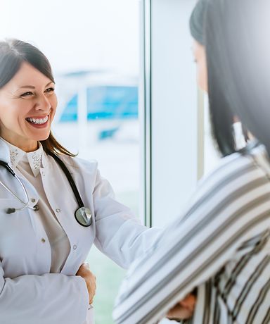 Eine Frau in einer gestreiften Bluse steht vor einer Ärztin. Die Ärztin lächelt die Frau an. 