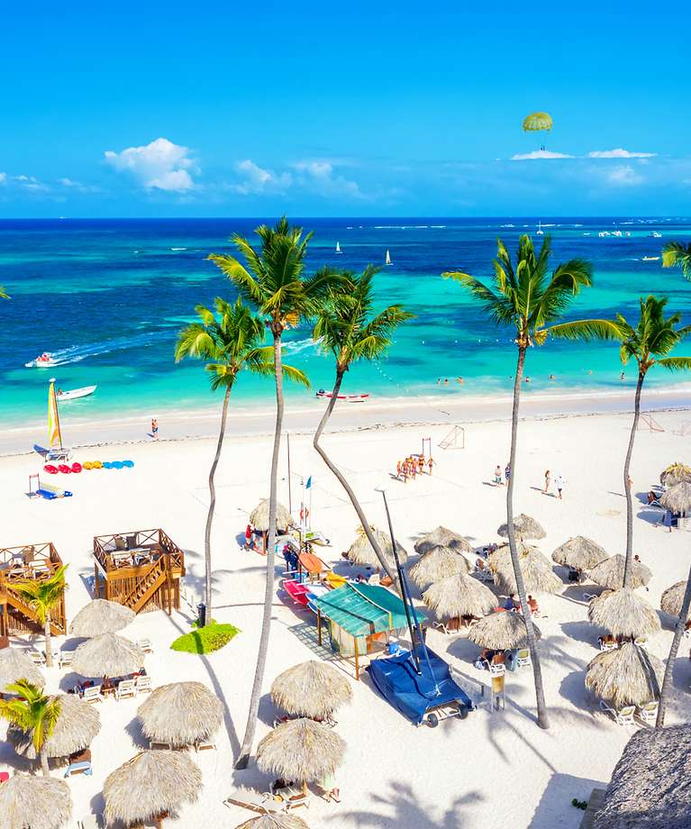 Man sieht bunte Häuser, die am Strand neben vieler Palmen stehen.