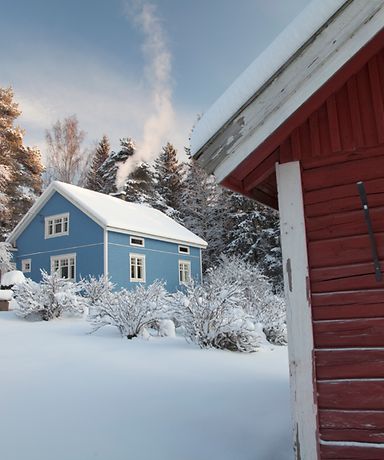 Man erkennt ein blaues Holzhaus umgeben von Schnee. Im Hintergrund sind schneebedeckte Bäume zu sehen.