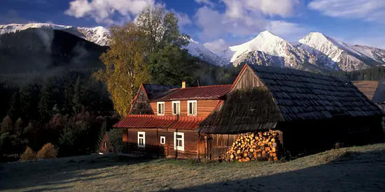 Man sieht eine Hütte und einen Holzschuppen am Fuße einer Berglandschaft.