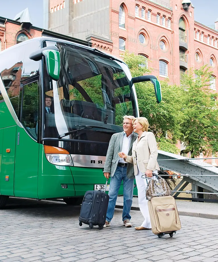 Zusehen sind zwei ältere Menschen, die mit ihren Koffern vor einem Bus stehen.