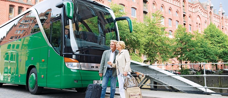 Zusehen sind zwei ältere Menschen, die mit ihren Koffern vor einem Bus stehen.