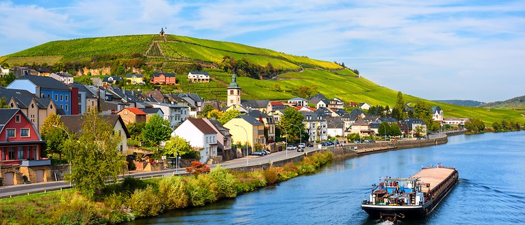 Man sieht ein Boot, dass auf der Mosel ist. Am Ufer stehen Häuser und es sind Weinberge zusehen.