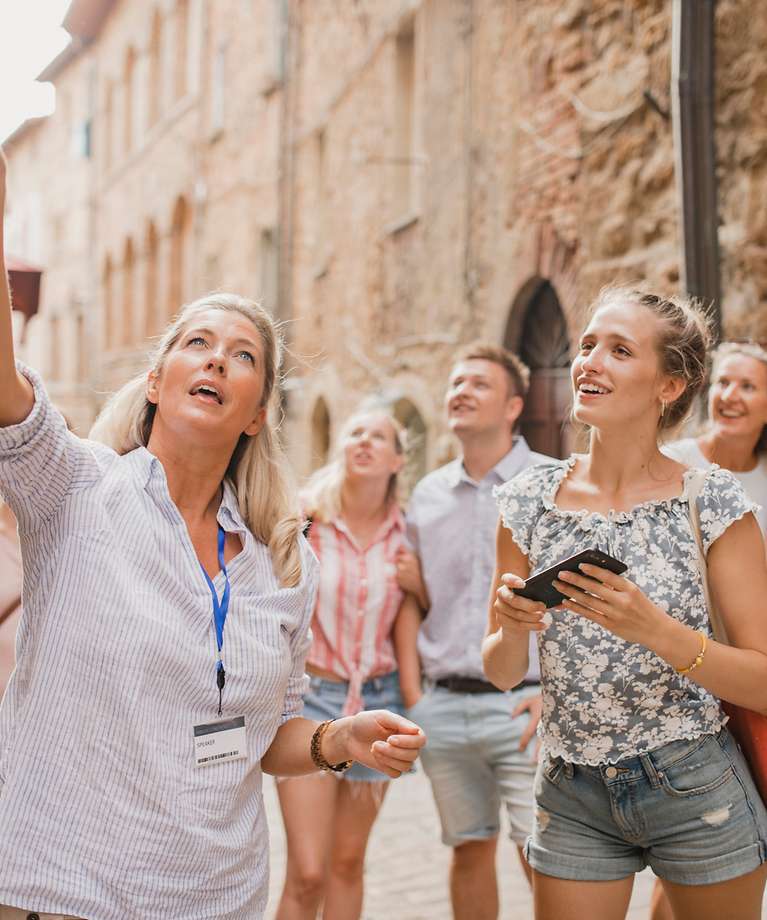 Eine Reisegruppe befindet sich in einer Altstadt. Eine Frau zeigt mit ihrem Finger auf ein Gebäude.