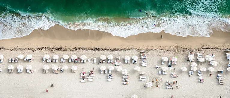 Man erkennt einen Strand von oben. Am Strand stehen viele Liegen und Sonnenschirme.