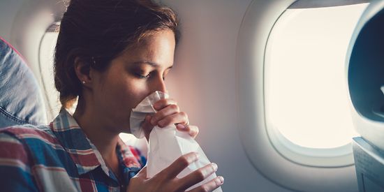 Eine Frau sitzt im Flugzeug. Vor ihrem Mund hält sie einen Spuckbeutel.