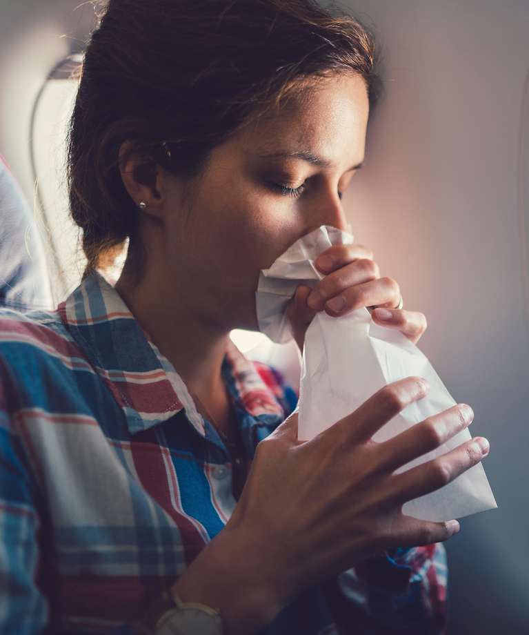 Eine Frau sitzt im Flugzeug. Vor ihrem Mund hält sie einen Spuckbeutel.