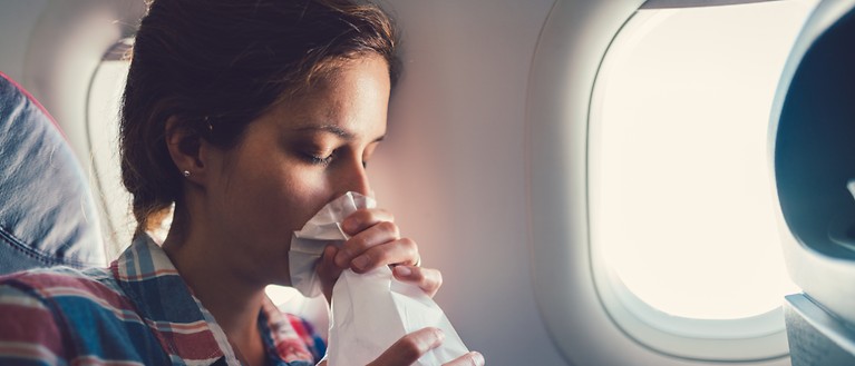 Eine Frau sitzt im Flugzeug. Vor ihrem Mund hält sie einen Spuckbeutel.