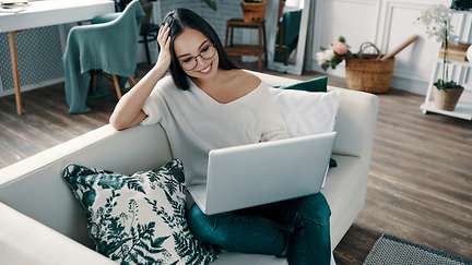 Eine Frau sitzt in ihrer Wohnung am Laptop und lächelt. 