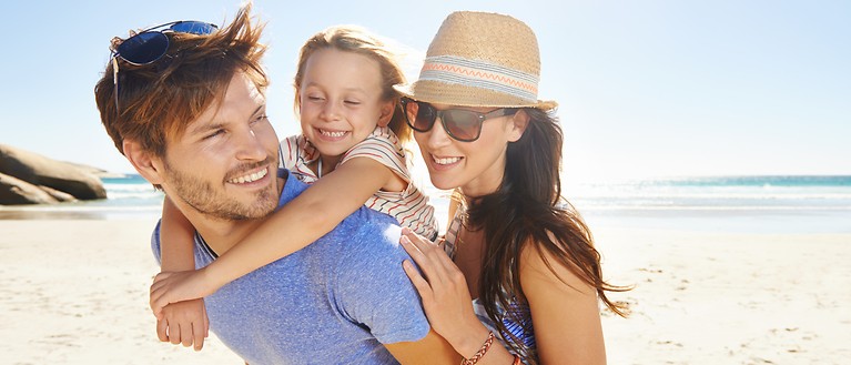 Mutter, Vater und deren Tochter sind am Strand. Im Hintergrund ist das Meer. 
