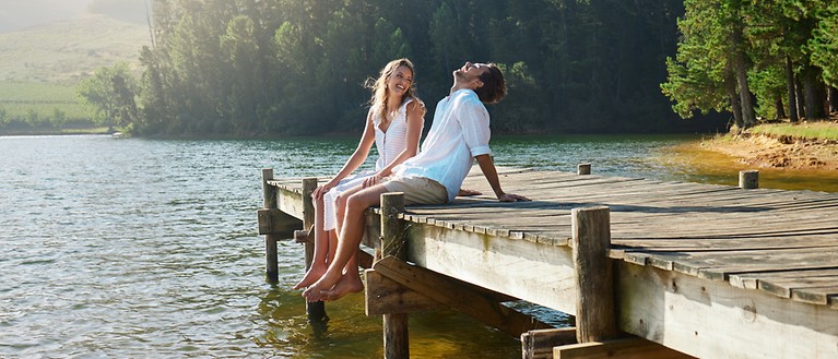 Ein Mann und eine Frau sitzen auf einem Steg, der ins Wasser hineinführt und lachen.