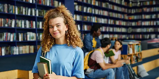 Eine junge Studentin steht mit einem Buch in der Hand in einer Bibliothek.