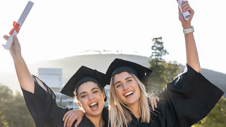 Abschlussfeier- Reisethemen Erasmus Man sieht zwei Studentinnen, die in Abschlusskleidung freudig ihr Zeugnisse in die Luft halten.