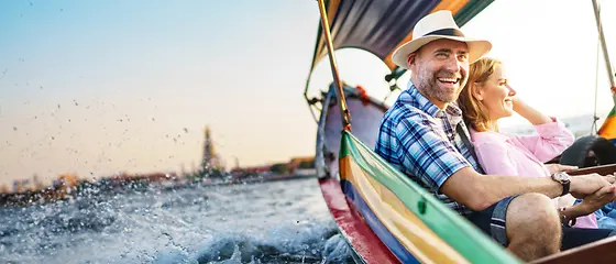 Ehepaar in einem Boot - Reiserücktrittsversicherung & Reiseabbruch-Versicherung Ein Ehepaar sitzt in einem bunten Boot. Im Hintergrund kann man Häuser sehen.