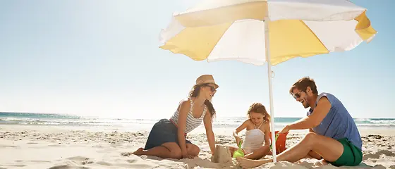 Eine Familie sitzt unter einem Sonnenschirm am Strand und spielen im Sand.