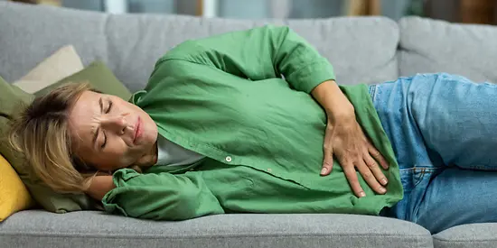 A woman in a green blouse is lying on the sofa, clutching her stomach in pain.