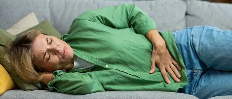 A woman in a green blouse is lying on the sofa, clutching her stomach in pain.