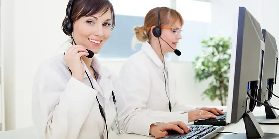 Two women wearing headsets are sitting at a computer, working.