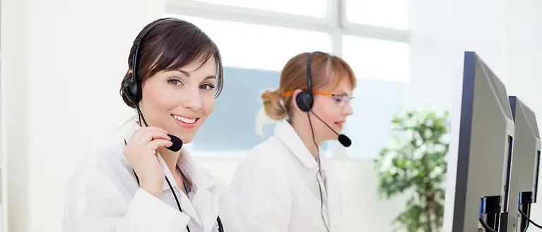 Two women wearing headsets are sitting at a computer, working.
